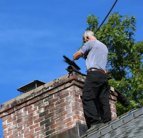 Person using a chimney brush beside a brick chimney on a sloped roof